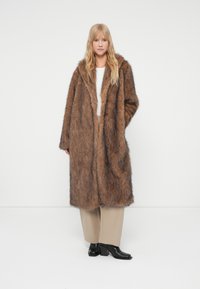 Cappotto in pelliccia sintetica marrone con cappuccio, caratterizzato da una consistenza morbida, vestibilità ampia e tasche laterali. Abbinato a pantaloni beige e stivali neri con tacco.