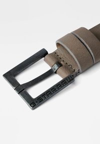 Brauner Leder-Gürtel mit einer rechteckigen Metallschnalle in dunklem Finish und geprägtem Logo. Sichtbare Steppnähte entlang des Leders.