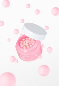 Lyserød krukke med små runde kapsler og hvid låg. Krukke omgivet af flydende lyserøde kugler på hvid baggrund.