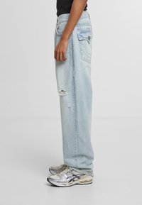 Jeans de mezclilla azul claro con un ajuste holgado, detalles desgastados y rasgaduras visibles, y diseño de cinco bolsillos. Combinados con zapatillas plateadas.