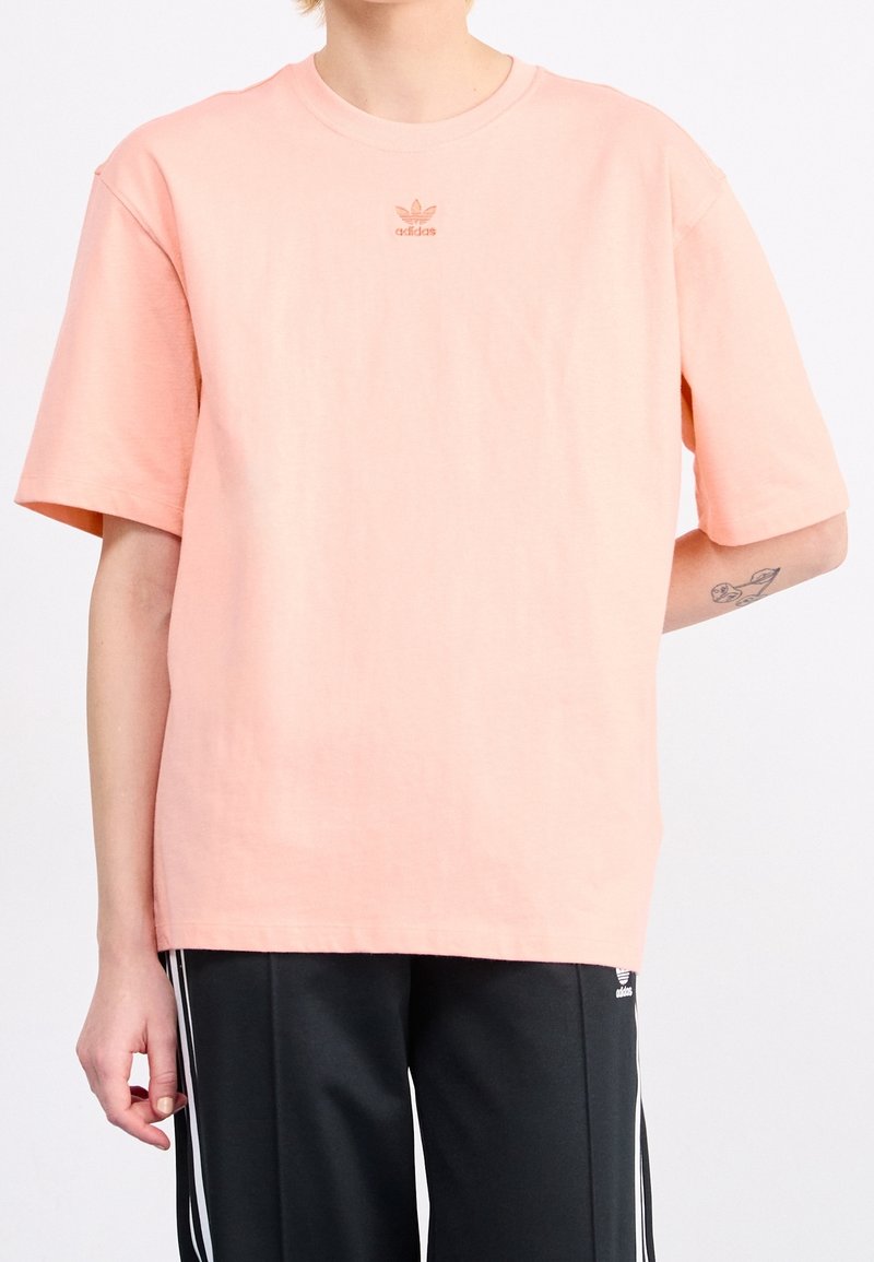 Osoba oblečená v světle růžovém tričku Adidas a černých teplácích Adidas s bílými pruhy, na levém předloktí je viditelné tetování.