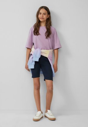 Paarse katoen t-shirt gecombineerd met marineblauwe fietsshorts. Een pastelkleurige gradient sweatshirt om de taille gewikkeld, en beige sneakers met witte accenten.