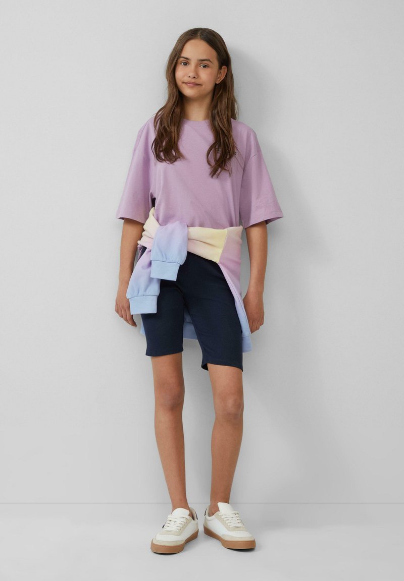 Paarse katoen t-shirt gecombineerd met marineblauwe fietsshorts. Een pastelkleurige gradient sweatshirt om de taille gewikkeld, en beige sneakers met witte accenten.