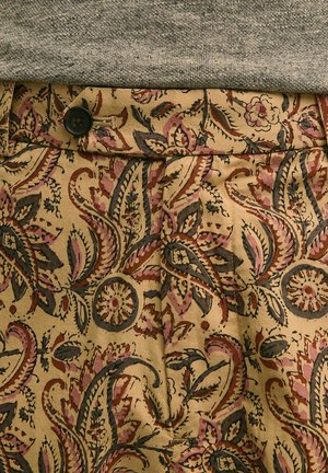 Pantalon beige à motifs floraux cachemire marron, rose et noir, maintenu par un bouton noir à la taille, associé à un haut en maille grise.