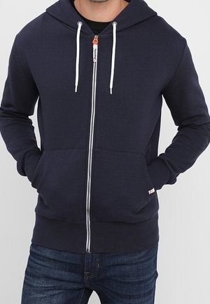 Mand iført en marineblå hættetrøje med lynlås med hvide snore og mørkeblå jeans, hænderne i hættetrøjens lommer.