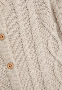 Cardigan beige lavorato a maglia con motivo a treccia e bottoni in legno sulla patta a coste.