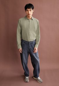 Olijfgroene langekout shirt met een voorzak, gecombineerd met losse donkere spijkerbroeken en grijze sneakers. Getextureerde stof, ingetogen stijl.