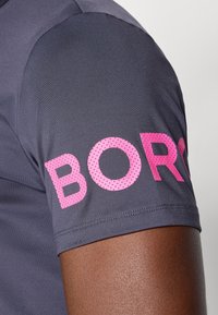 Pelēks sporta krekls ar gludu tekstūru, uz kura īsajām piedurknēm ir drosmīgs rozā "BORN" teksts. Šuves redzamas netālu no apakšmalas.