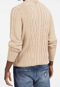 Beiger Strickpullover mit Zopfmuster, strukturiertem Design, gerippten Bündchen und einem Stehkragen, kombiniert mit blauen Jeans aus Denim.