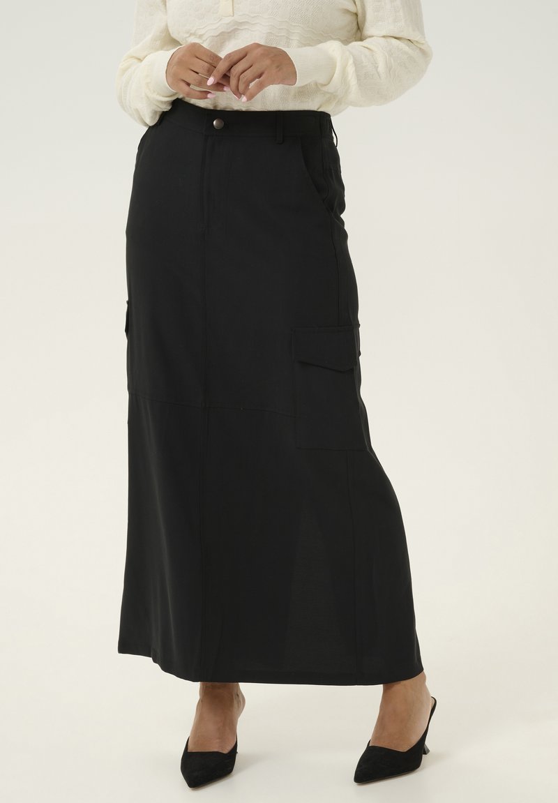 Zwarte maxi rok met een rechte snit, voorzien van zijzakken, een knoopsluiting en een gladde textuur. Gecombineerd met puntige zwarte hakken.