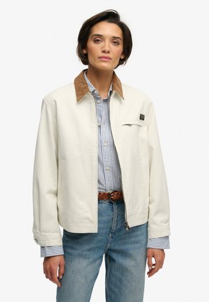 Kvinde iført en cremejakke med brun krave over en blåstribet skjorte og jeans, stående mod en hvid baggrund.