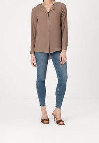 Blouse marron à manches longues avec un col, en tissu léger, devant boutonné, associée à un jean skinny bleu et des sandales à talons marron.