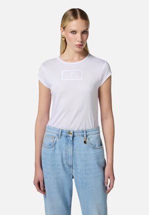 T-shirt en coton blanc à manches courtes et à encolure ronde, avec un logo rectangulaire texturé au centre. Associé à un jean en denim bleu clair.
