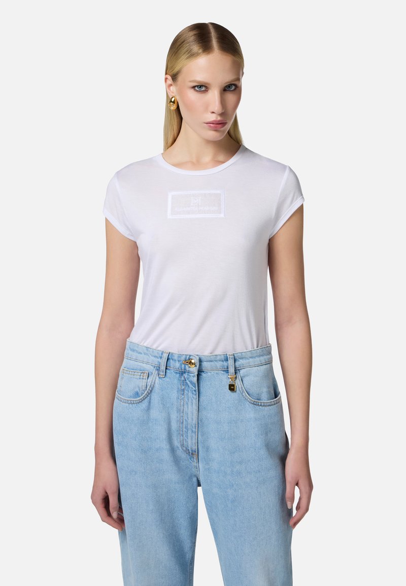 Camiseta de algodón blanca de manga corta y escote redondo, con un logotipo rectangular texturizado en el centro. Combinada con unos jeans de mezclilla azul claro.