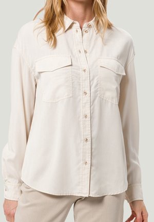 Mujer que lleva una camisa beige de manga larga con botones y dos bolsillos con solapa en el pecho, junto con pantalones beige.