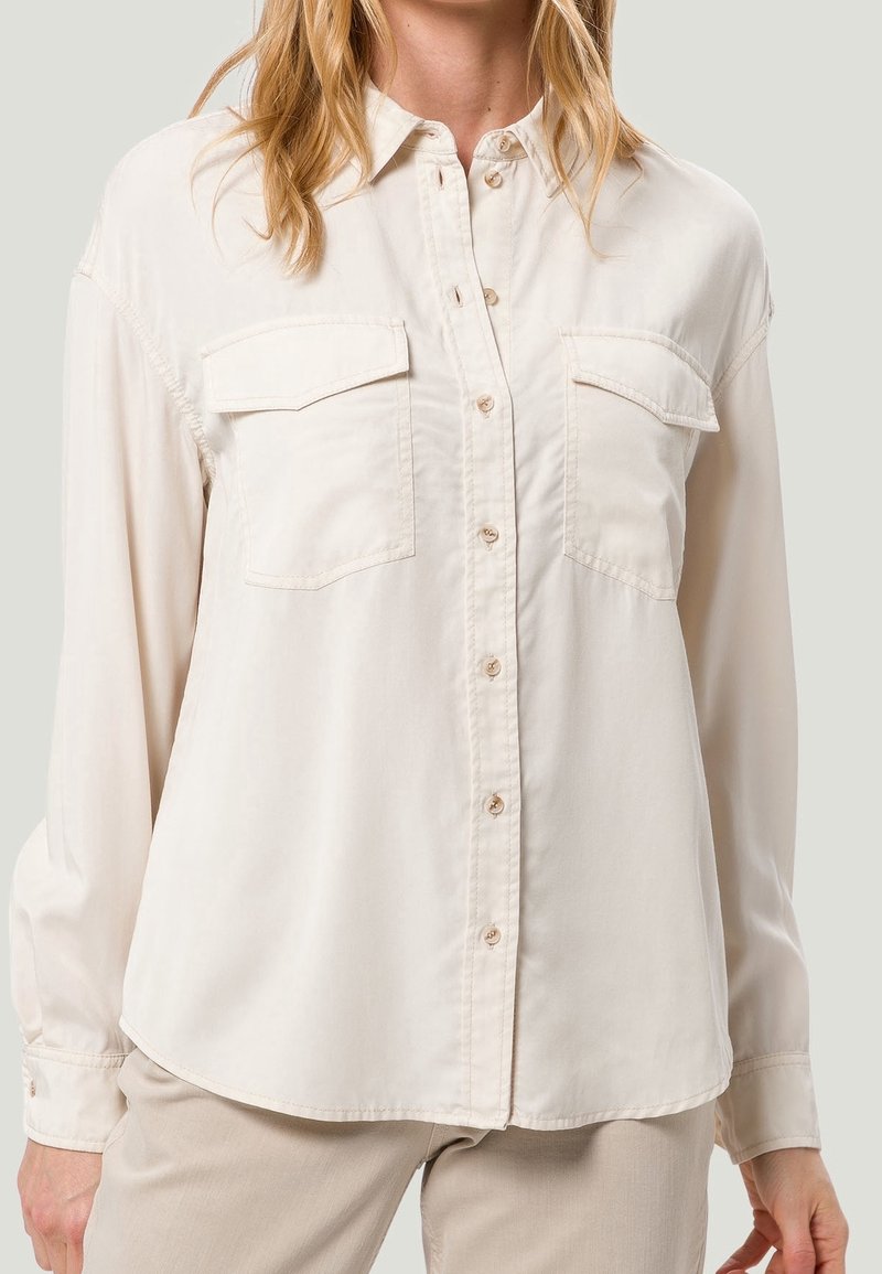 Femme portant une chemise beige à manches longues, boutonnée, avec deux poches à rabat sur la poitrine, et un pantalon beige.