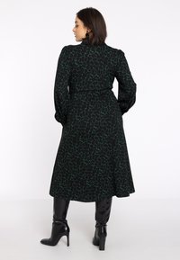 Robe noire avec motif abstrait vert, dotée de manches longues et d'une taille ceinturée. Associée à des bottes à talons hauts noires.