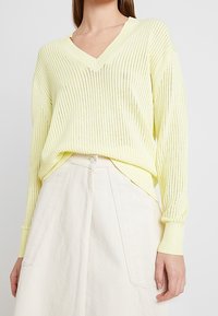 Jasnożółty, dziergany pulower z dekoltem w serek i prążkowaną fakturą, zestawiony z białą spódnicą o kroju litery A z zapięciem na guziki z tyłu.