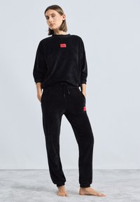 Čierny sametový loungewear set, ktorý pozostáva z mikiny s okrúhlym výstrihom a nohavíc na šnúrku, oboje so značkovou červenou nášivkou. Mäkká textúra, uvoľnený strih.