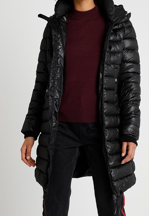 Doudoune noire avec capuche, présentant des sections matelassées verticales, une fermeture à boutons-pression et des poignets côtelés. Portée par-dessus un pull bordeaux.