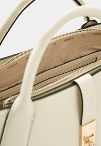 Sac à main couleur crème avec une surface texturée, des accessoires en or et des bords noirs contrastants. L'intérieur est doublé de tissu avec le logo de la marque.