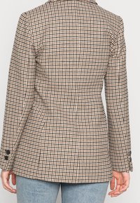 Brązowy i kremowy marynarka w wzór houndstooth z dopasowanym krojem, klapkami oraz dwoma czarnymi guzikami na każdym rękawie. Teksturowana tkanina.