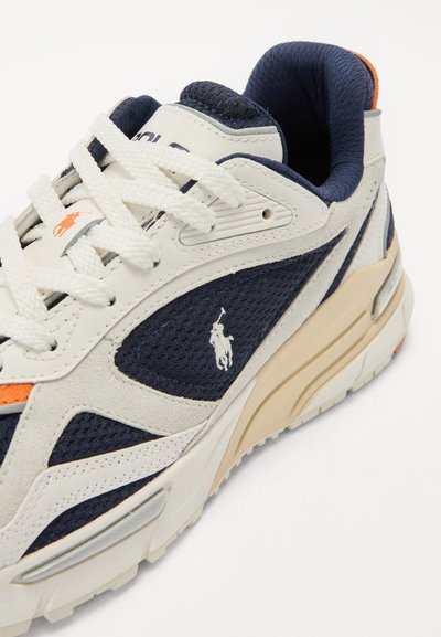 Ténis com uma combinação de malha azul-marinho e camurça bege, com atacadores brancos e um detalhe de logótipo. Amortecimento visível no design da sola.