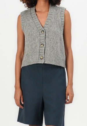 Personne portant un gilet gris tricoté sans manches avec des boutons, associé à un short sombre taille haute, sur fond blanc uni.