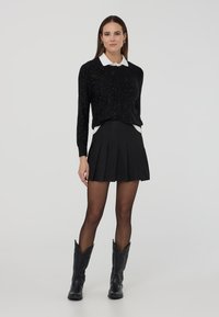 Donna con un cardigan nero a pois, camicia bianca con colletto, gonna mini plissettata nera, collant trasparenti e stivali da cowboy neri, in piedi di fronte a uno sfondo semplice.