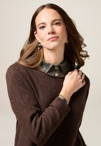 Brauner gestrickter Pullover mit subtilen Glitzereffekten, kombiniert mit einem blumenbedruckten Hemd mit Kragen. Die Haare sind wellig und die Ohrringe haben farbenfrohe Akzente.