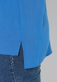 Camicia blu reale con orlo leggermente curvo e spacchi laterali, realizzata in un tessuto leggero. Abbinata a jeans in denim scuro.