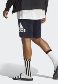 Námornícke modré športové šortky s bielym logom Adidas, spolu s čierno-bielymi pruhovanými ponožkami a čiernymi šľapkami. Hladká tkanina, ležérny dizajn.