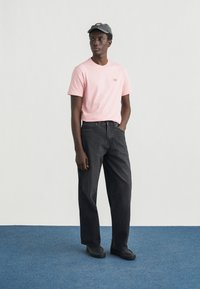 Ροζ βαμβακερό t-shirt με μικρό λογότυπο, συνδυασμένο με μαύρο τζιν παντελόνι. Το μοντέλο φοράει σκούφο σκούρου χρώματος και μαύρα παπούτσια, στέκεται σε μπλε χαλί.