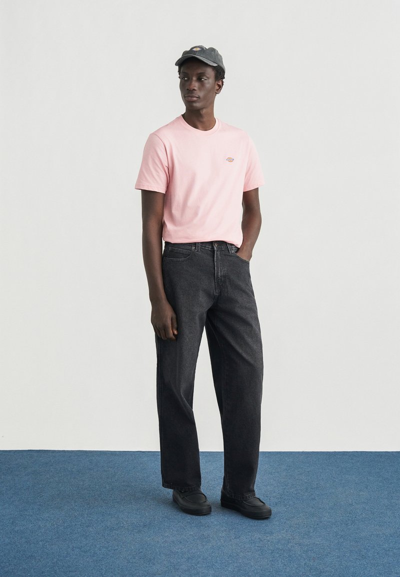 Roze katoenen T-shirt met een klein logo, gecombineerd met zwarte spijkerbroek. Model draagt een donkere pet en zwarte schoenen, staand op een blauwe vloer.