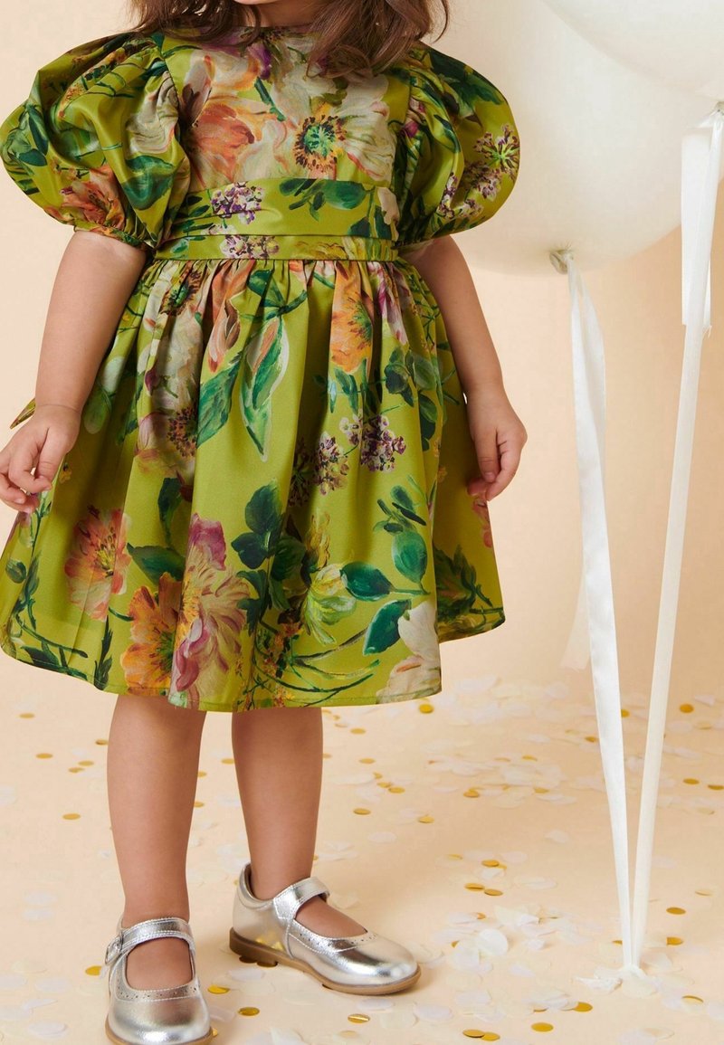 Enfant portant une robe fleurie verte avec des manches bouffantes et des chaussures Mary Jane argentées, debout parmi des confettis blancs et dorés près de ballons pâles.