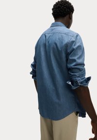 Camicia in denim di un blu chiaro con colletto button-down, maniche lunghe arrotolate ai polsini e orlo curvo. Texture morbida con una finitura sbiadita.
