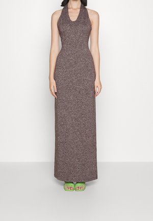 Vrouw draagt een mouwloze, bruine halternek maxi-jurk gecombineerd met felgroene sandalen met vierkante neus, staand tegen een witte muur.