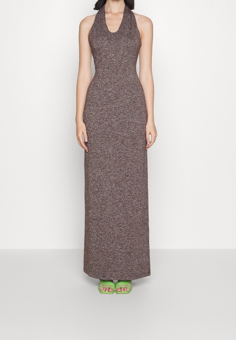 Femme portant une robe maxi marron sans manches à col licou, associée à des sandales vert vif à bout carré, debout contre un mur blanc.