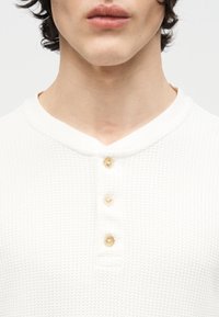 Νεαρός που φοράει λευκό υφασμάτινο πουκάμισο henley με τρία μπεζ κουμπιά, εμφανιζόμενος από τον λαιμό έως το πάνω μέρος του στήθους.