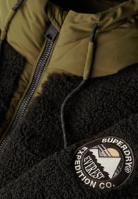 Zwarte fuzzy fleece jas met een olijfgroene capuchon, voorzien van een ritsluiting en een rond logopatch op de linker mouw.