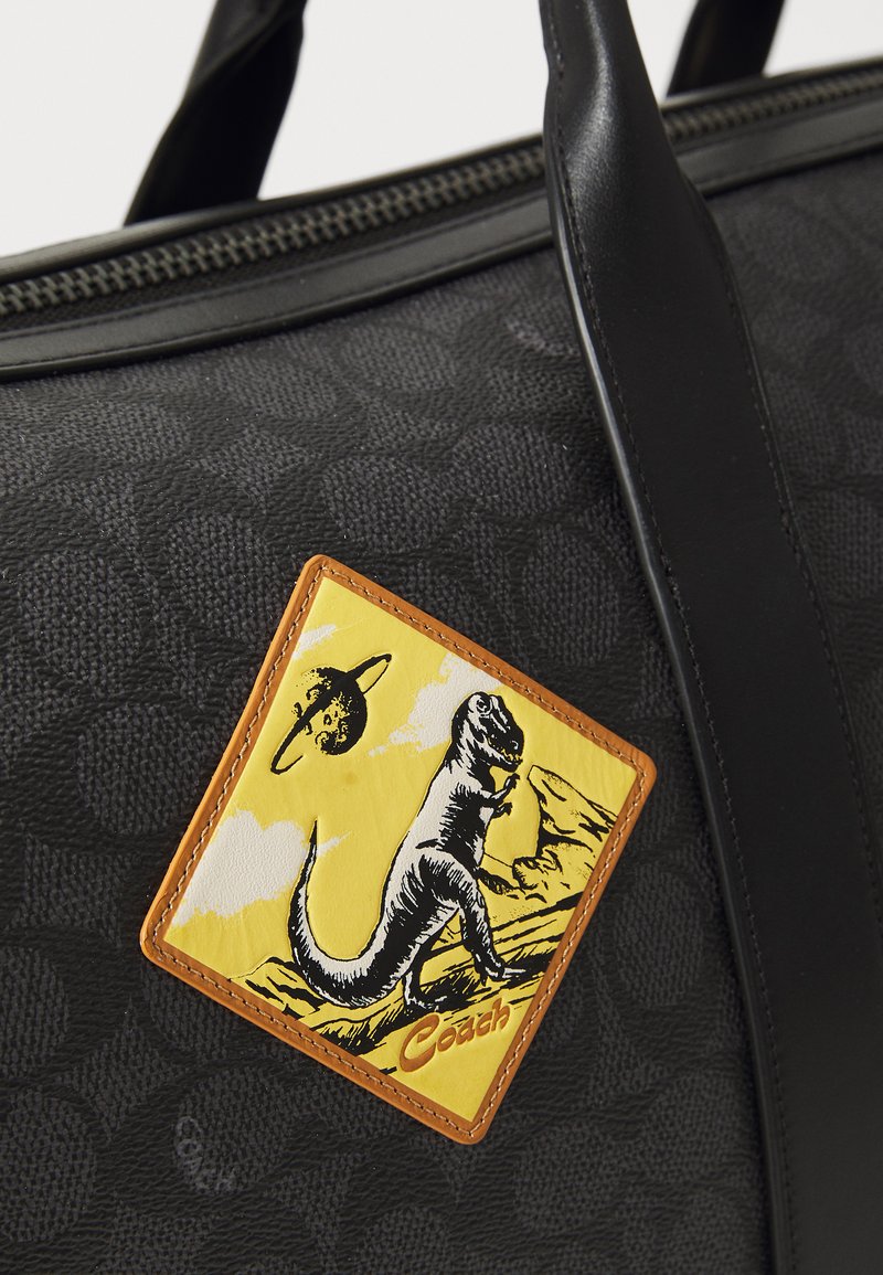 Black patterned fabric bag with smooth black leather accents. Features an orange rectangular patch with a dinosaur illustration on a yellow background.