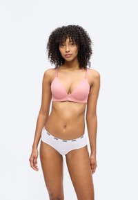 Γυναίκα με σγουρά μαλλιά που φοράει ροζ σουτιέν και λευκά εσώρουχα Puma, στέκεται μπροστά σε απλό φωτεινό φόντο.