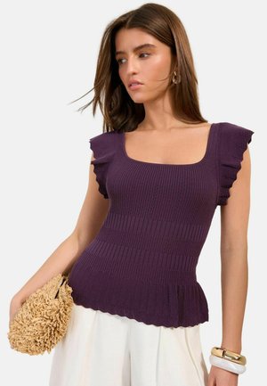 Noor naine, kes kannab keha järgivat lilla kudumit rüüsidega varrukatel ja ääres, hoides beeži tekstuuriga käekotti, valge taustaga.