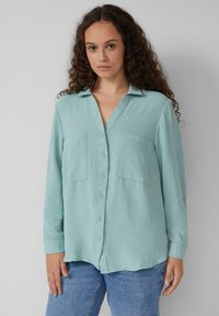 Helles blaues Hemd mit V-Ausschnitt, zwei vorderen Taschen, langem, hochgekrempeltem Ärmel und einer strukturierten Stoffoberfläche.