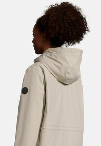 Beige waterdichte jas met een capuchon, voorzien van een gestructureerde kraag, een ronde stoffen logo patch op de mouw en een ruime pasvorm.