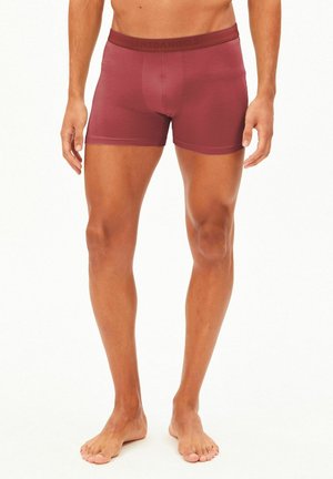 Mannelijk model draagt bordeaux boxershort, staat blootsvoets met ontspannen handen langs het lichaam tegen een effen achtergrond.