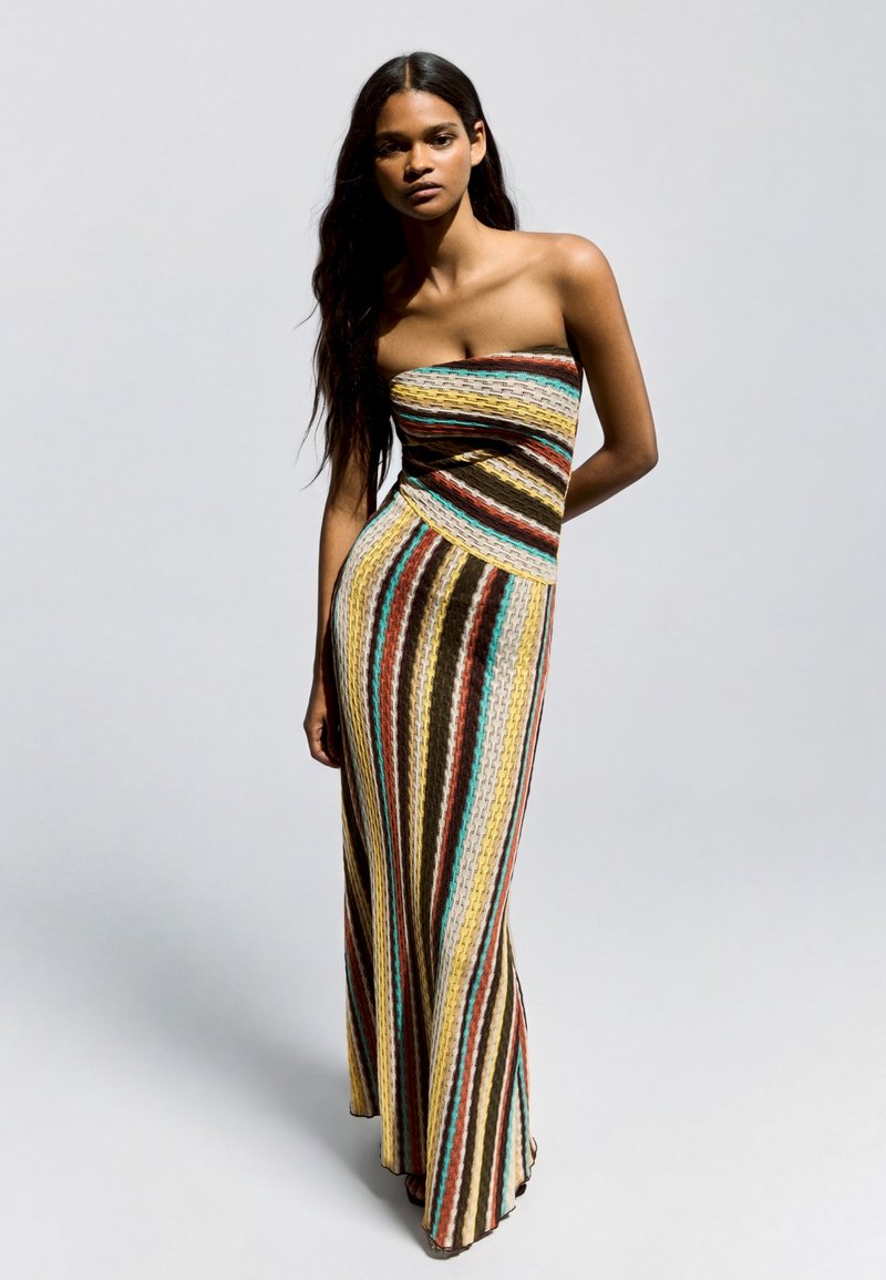 Femme aux longs cheveux foncés portant une robe longue sans bretelles avec des rayures verticales multicolores, debout devant un fond clair uni.