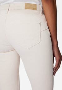 Jean en denim rose clair avec une coupe ajustée, doté d'un patch en cuir camel à la taille, de poches arrière brodées, et d'une texture subtile.