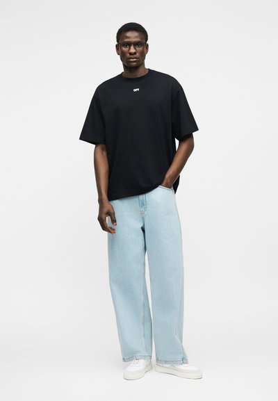 Άνδρας φοράει μαύρο υπερμεγέθες T-shirt με λογότυπο "OFF", ανοιχτό μπλε τζιν παντελόνι με φαρδιές ποδιές, λευκά αθλητικά παπούτσια και γυαλιά, στέκεται μπροστά σε απλό φόντο.