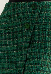 Jupe en tweed vert présentant un motif texturé en grille, avec des fils dorés et deux boutons verts à la taille pour la fermeture.